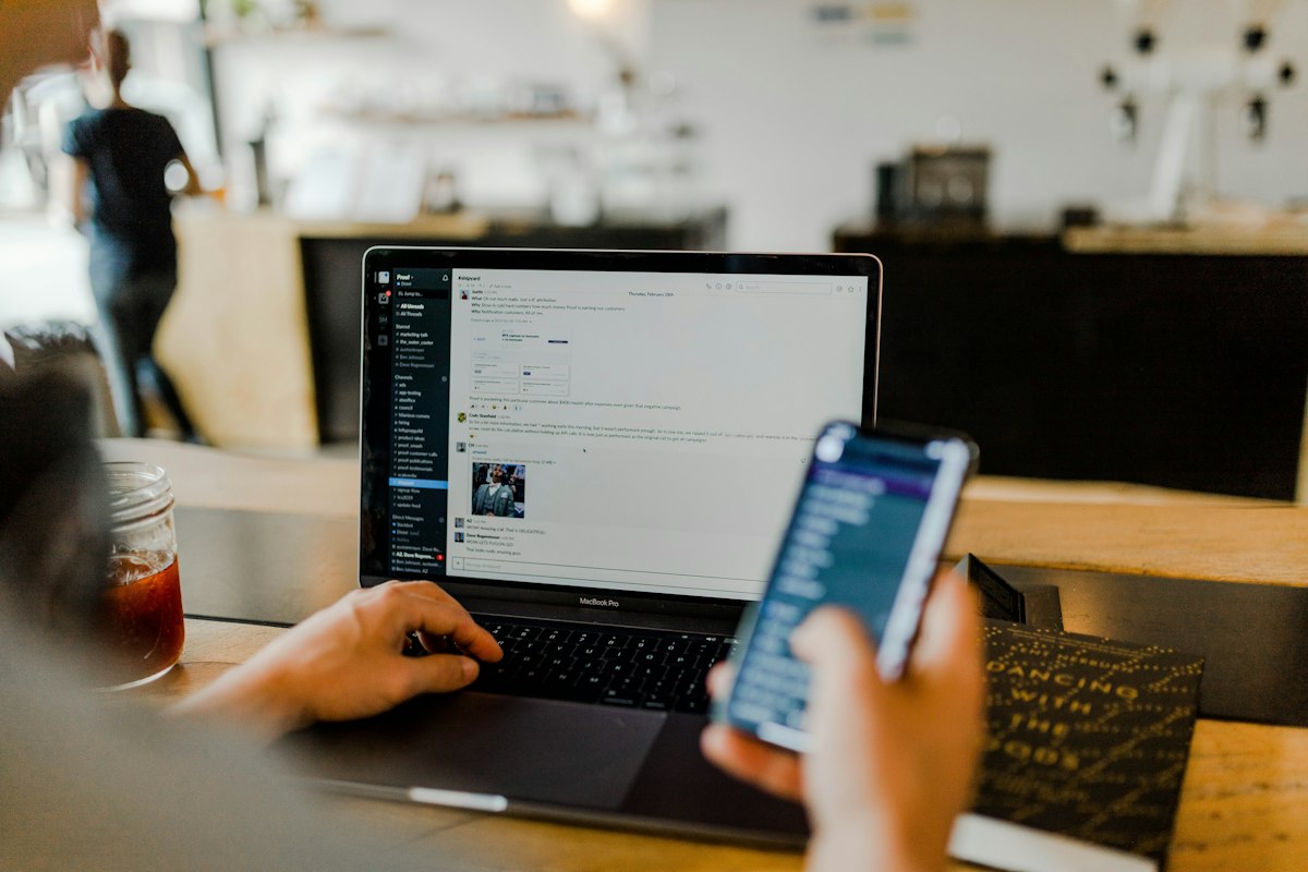 Someone typing on a laptop with a chat app open while holding a phone in a cafe